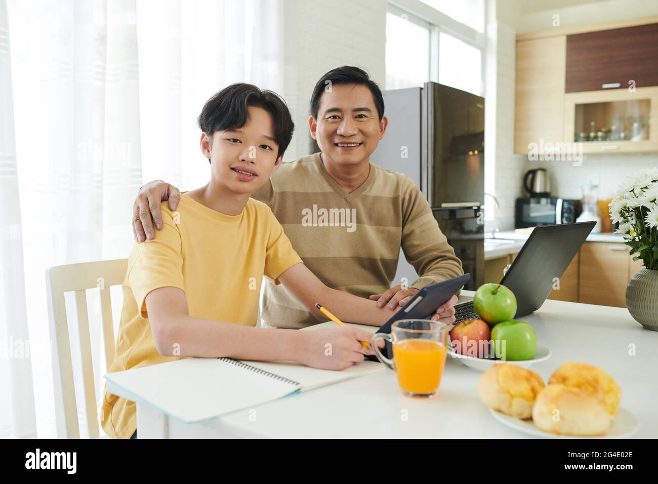 Porträt eines glücklichen umarmenden Vaters und Sohnes, der beim Arbeiten und Hausaufgaben am Küchentisch sitzt Stockfoto