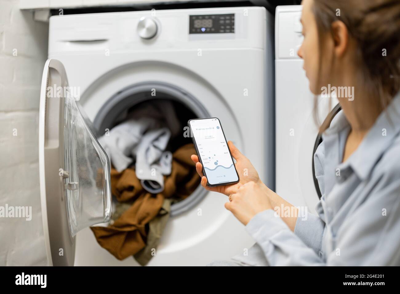 Die Frau steuert die Waschmaschine mit einem Smartphone Stockfoto
