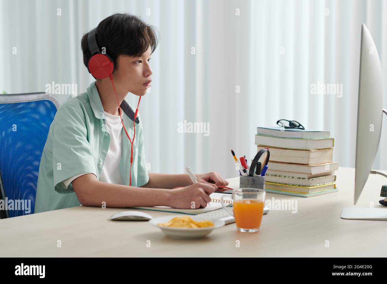Konzentrierter seriöser Schuljunge in Kopfhörern, der Lernvideos auf dem Computer ansieht und in Notizblock schreibt Stockfoto