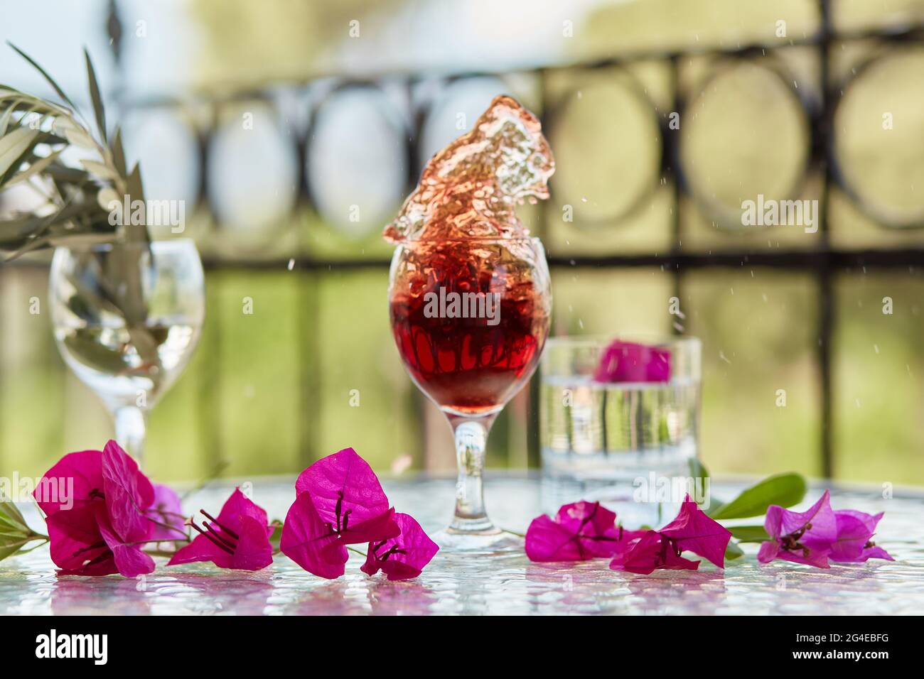 Elegantes und farbenfrohes Glas Rotwein, Wein spritzt ins Glas. Im Sommer helle Cocktails auf dem Hintergrund. Dekorativ rosa Blumen und der Zweig a Stockfoto