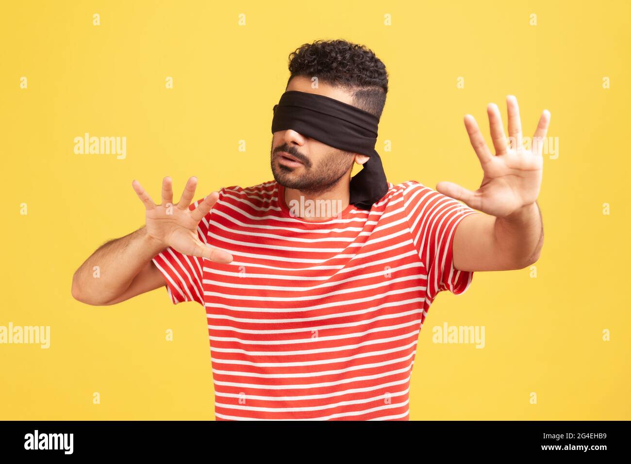 Frustrierter bärtiger Mann in gestreiftem T-Shirt mit Augenbinde, der versucht, sich in der Dunkelheit zu bewegen und den Raum mit den Händen zu berühren, Sehprobleme. Stu Stockfoto