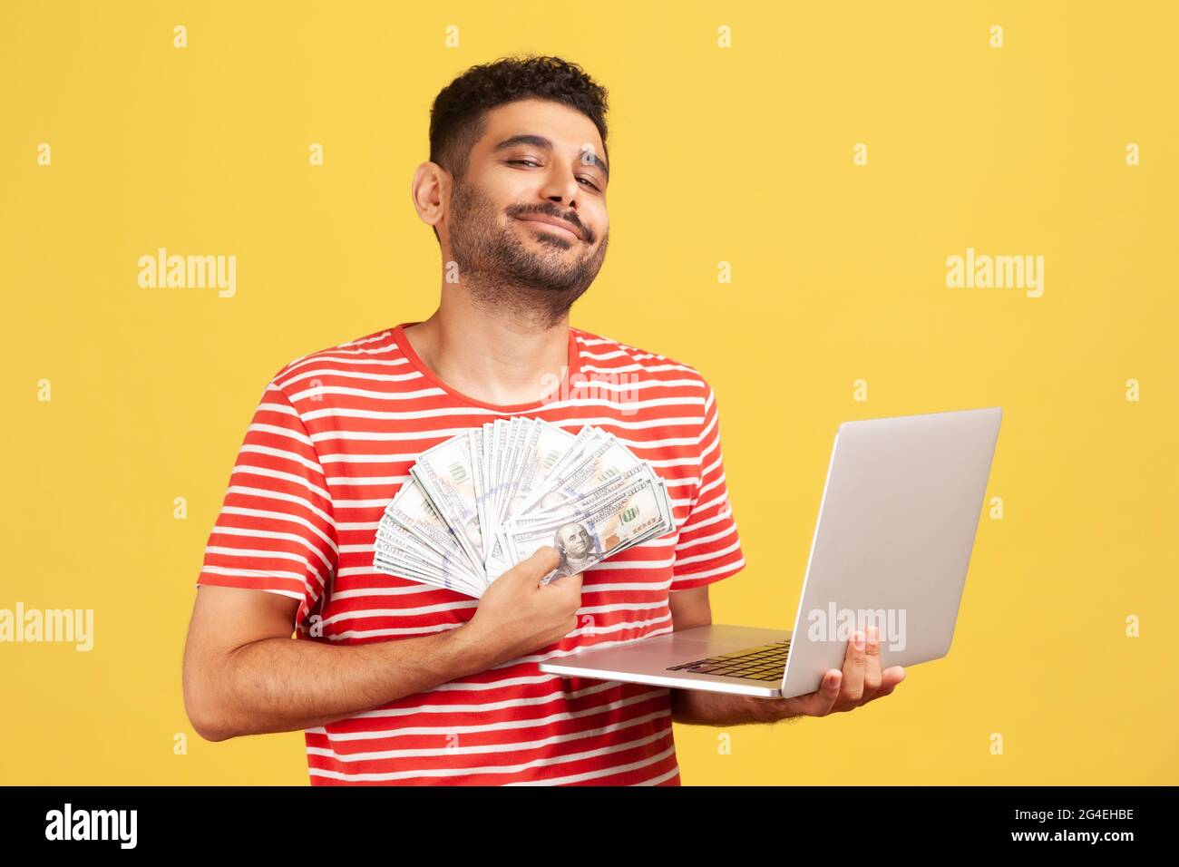 Selbstbewusster Mann, der lächelnd in den Händen hält, Laptop und Fan von hundert Rechnungen, stolz auf sein Gehalt, das er online arbeitet, Lotteriesieg, Wetten. Stockfoto