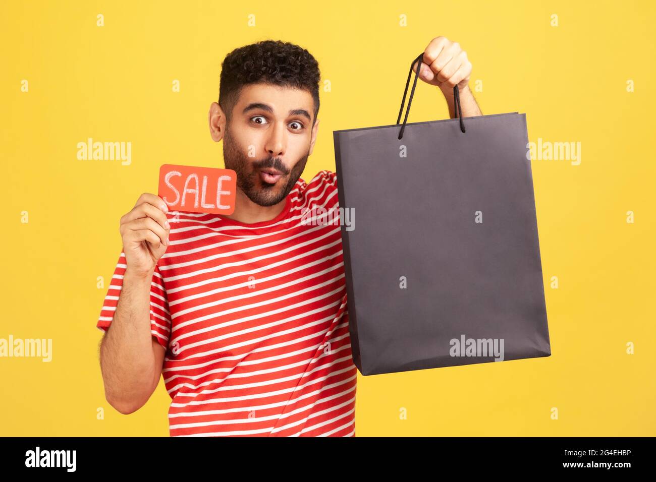 Überrascht bärtiger Mann, der mit Erstaunen die Kamera ansah und die Karte mit Verkaufsaufschrift und Einkaufstasche in den Händen hielt, schockiert über Rabatte. Innen Stockfoto