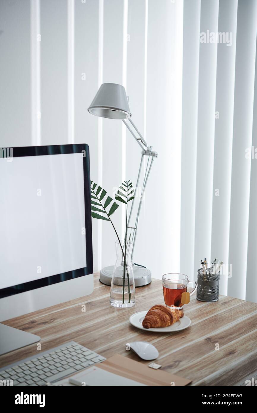 Tasse Tee und frisches Croissant auf dem Schreibtisch des Heimbüros neben Computer, Lampe und Vase mit Blättern Stockfoto
