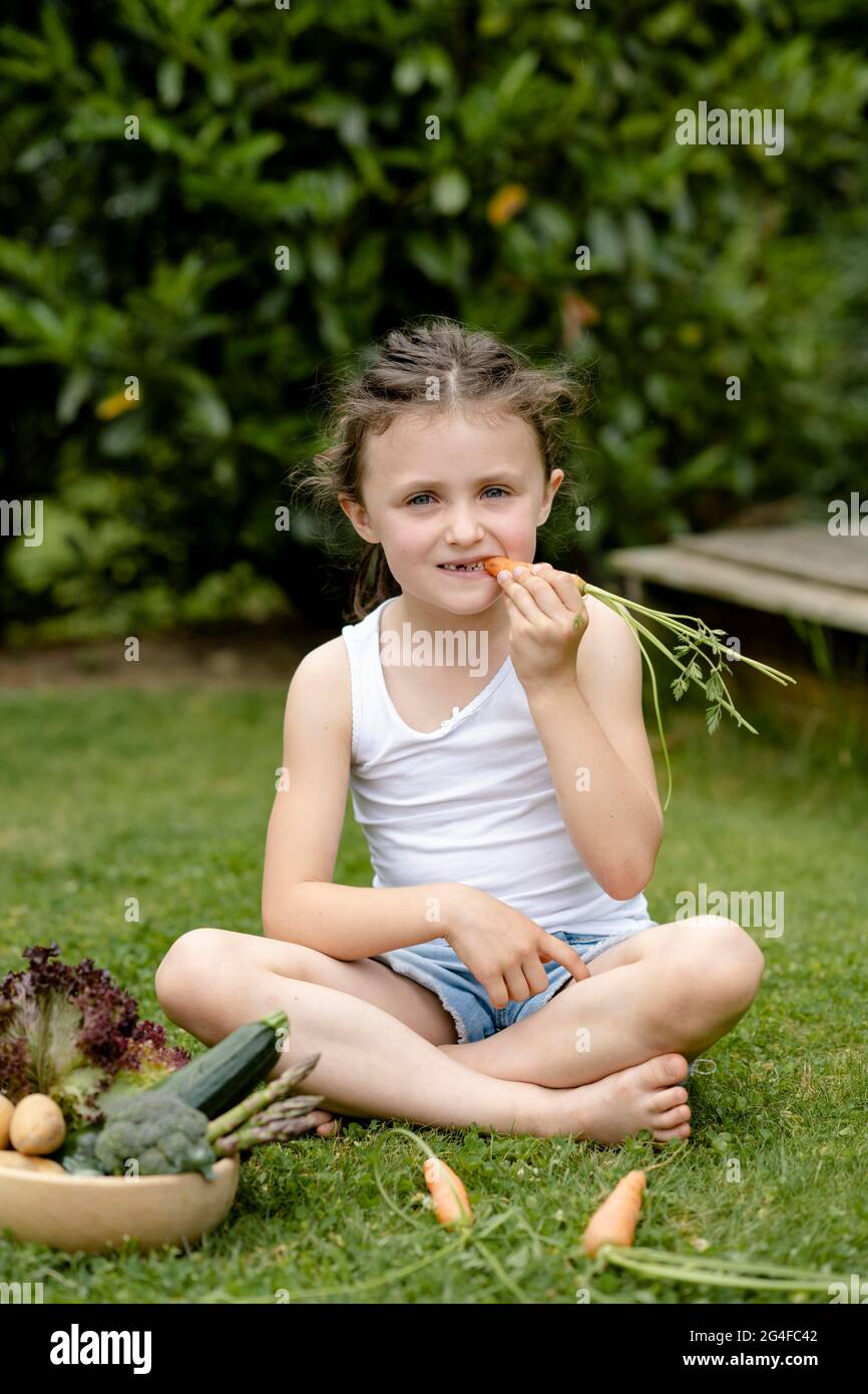 Mädchen mit Gemüskorb sitzt auf der Wiese und beißt in eine Karotte Stockfoto