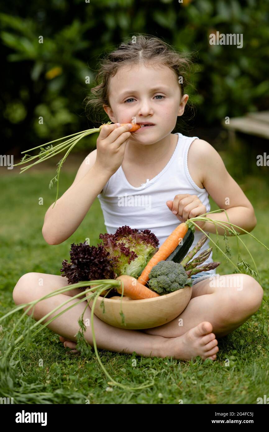 Mädchen mit Gemüskorb sitzt auf der Wiese und beißt in eine Karotte Stockfoto