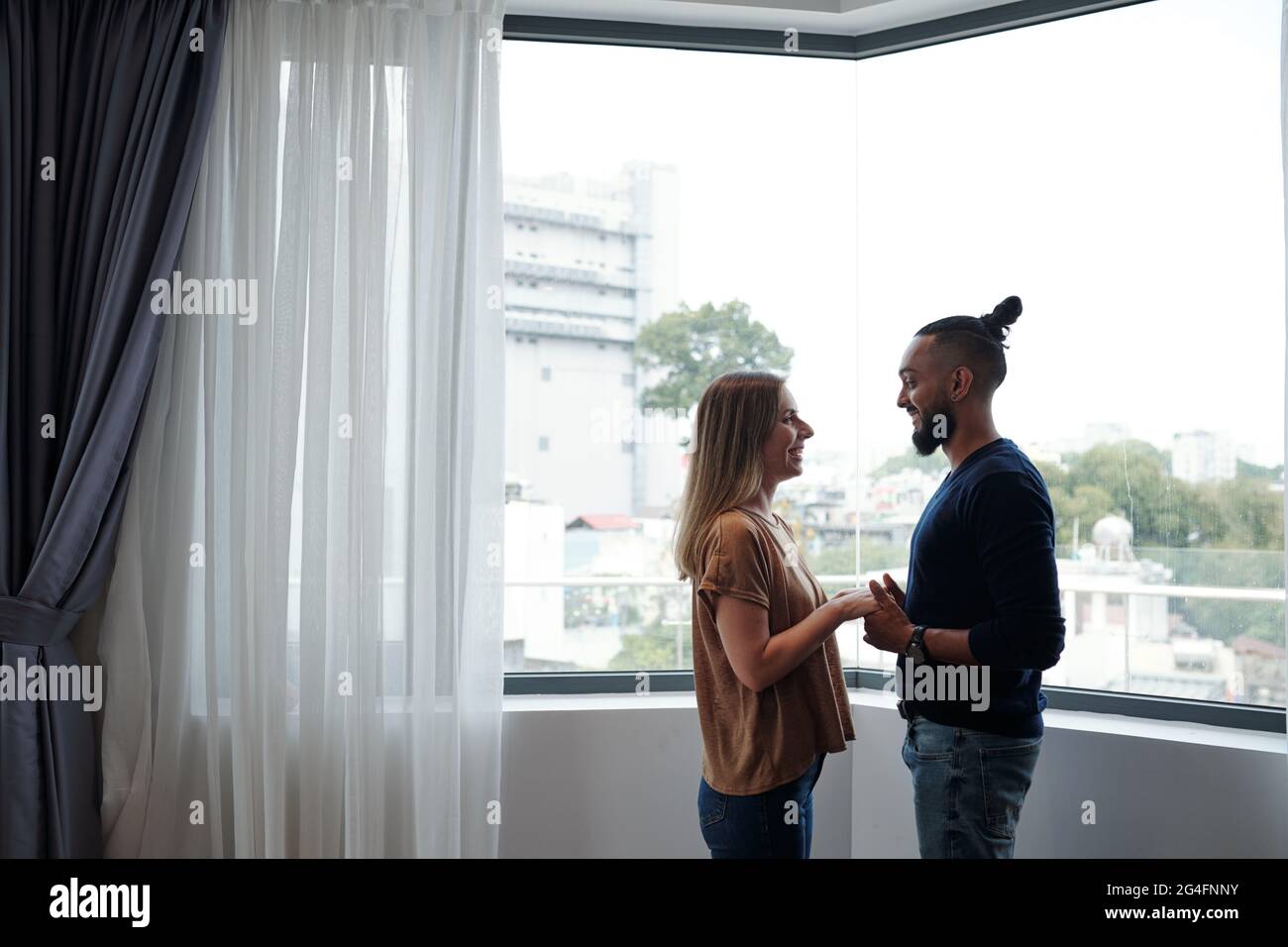 Glücklicher Freund und Freundin halten sich die Hände und schauen einander an, wenn sie gegen ein großes Wohnungsfenster stehen Stockfoto