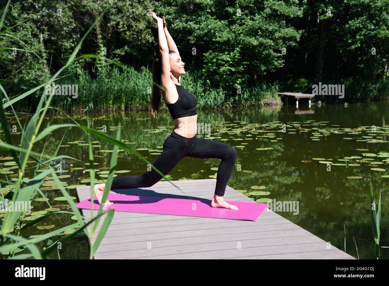 Frau in den Dreißigern praktiziert Yoga und Meditation. Fit Frau auf Holzplattform am Teich, umgeben von Bäumen und Wasser. Stockfoto