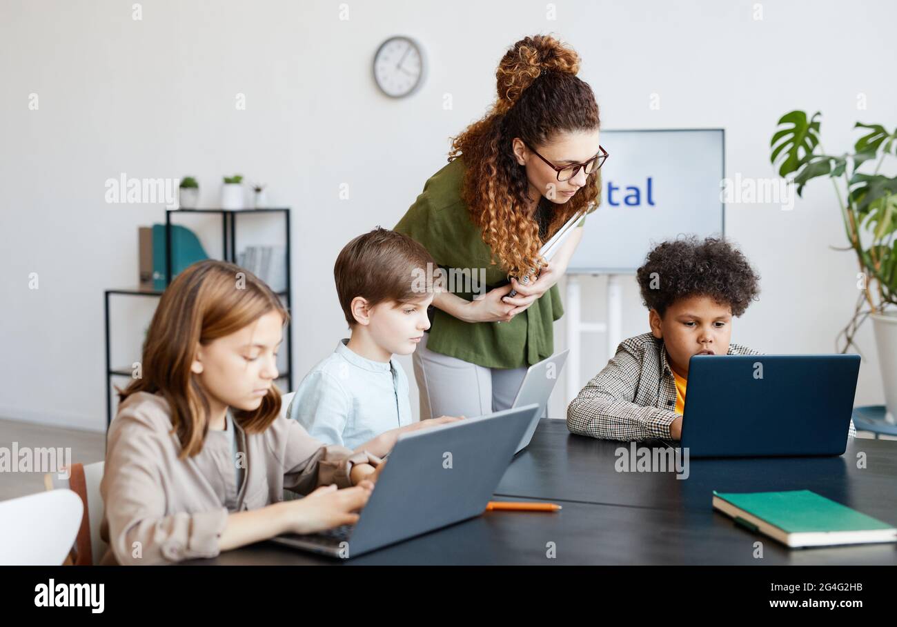 Verschiedene Gruppen von Kindern, die während DES IT-Unterrichts in der Schule mit einer jungen Lehrerin Computer benutzen Stockfoto