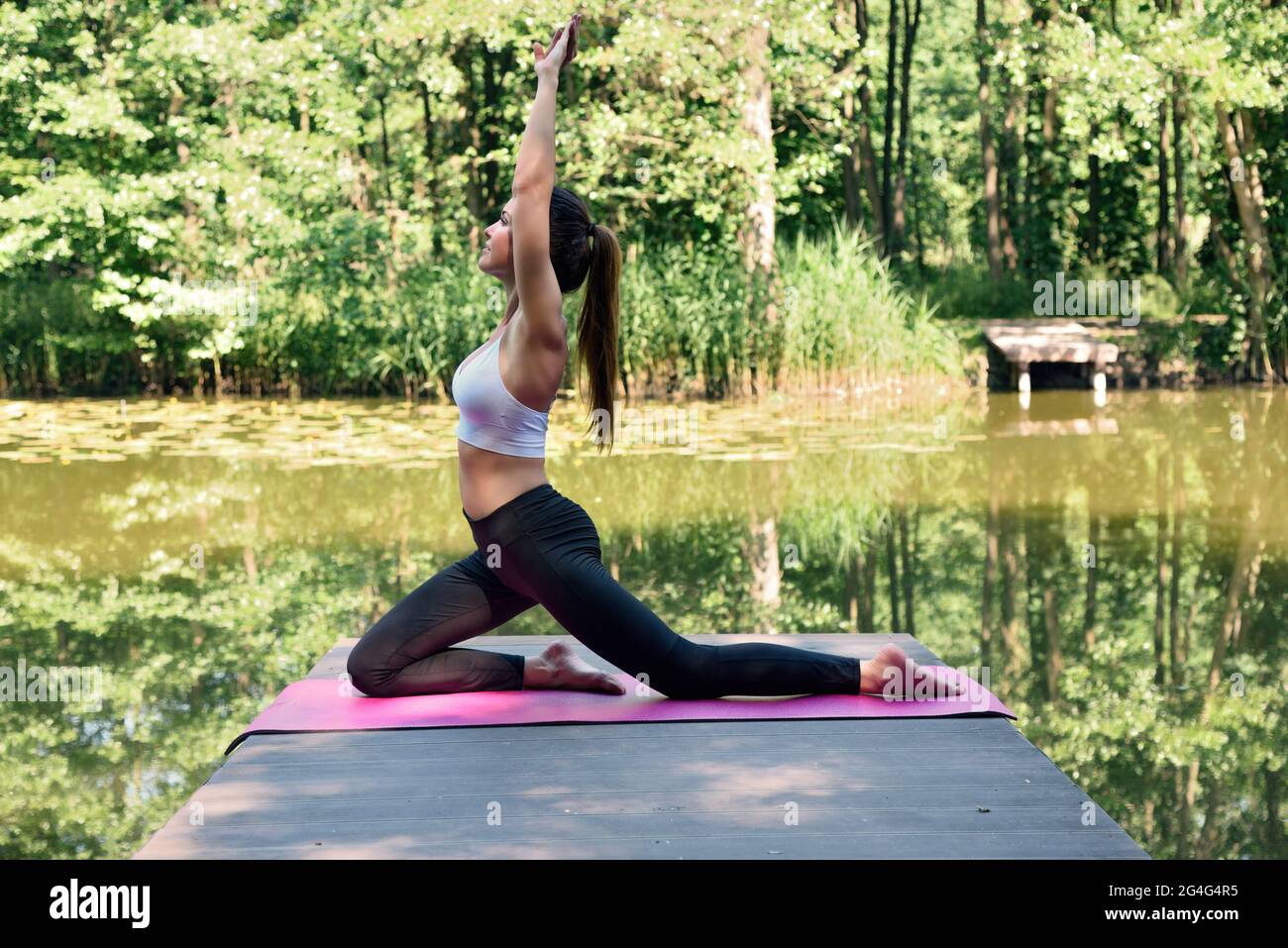 Frau in den Dreißigern praktiziert Yoga und Meditation. Fit Frau auf Holzplattform am Teich, umgeben von Bäumen und Wasser. Stockfoto