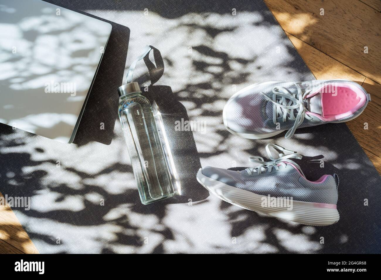 Laptop, Sneaker und Wasserflasche auf grauem Yogamatte-Hintergrund mit direkter Sonneneinstrahlung und Schatten. Online-Fitness-Programm, Home Workout. Workout Stockfoto