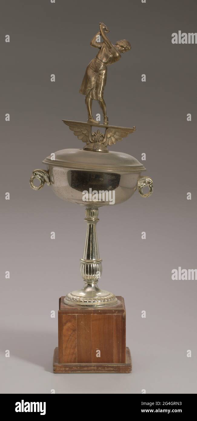 Eine Trophäe der United Golf Association "Amateur Women Championship Flight Winners", die 1968 von der afroamerikanischen Golferin Ethel Funches gewonnen wurde. Die Trophäe besteht aus Metall, wahrscheinlich einer Legierung, die wie Gold lackiert ist. Es liegt auf einer 5" dunkelbraunen quadratischen Holzbasis, die auf einer 3/4" dicken flachen Holzbasis liegt. Die Trophäe ist im Gold-Cup-Stil gehalten. Es gibt eine kreisförmige Basis mit einer strukturierten Kante. Die Säule ist unten abgerundet, wird oben dünner und endet kugelförmig. Stockfoto