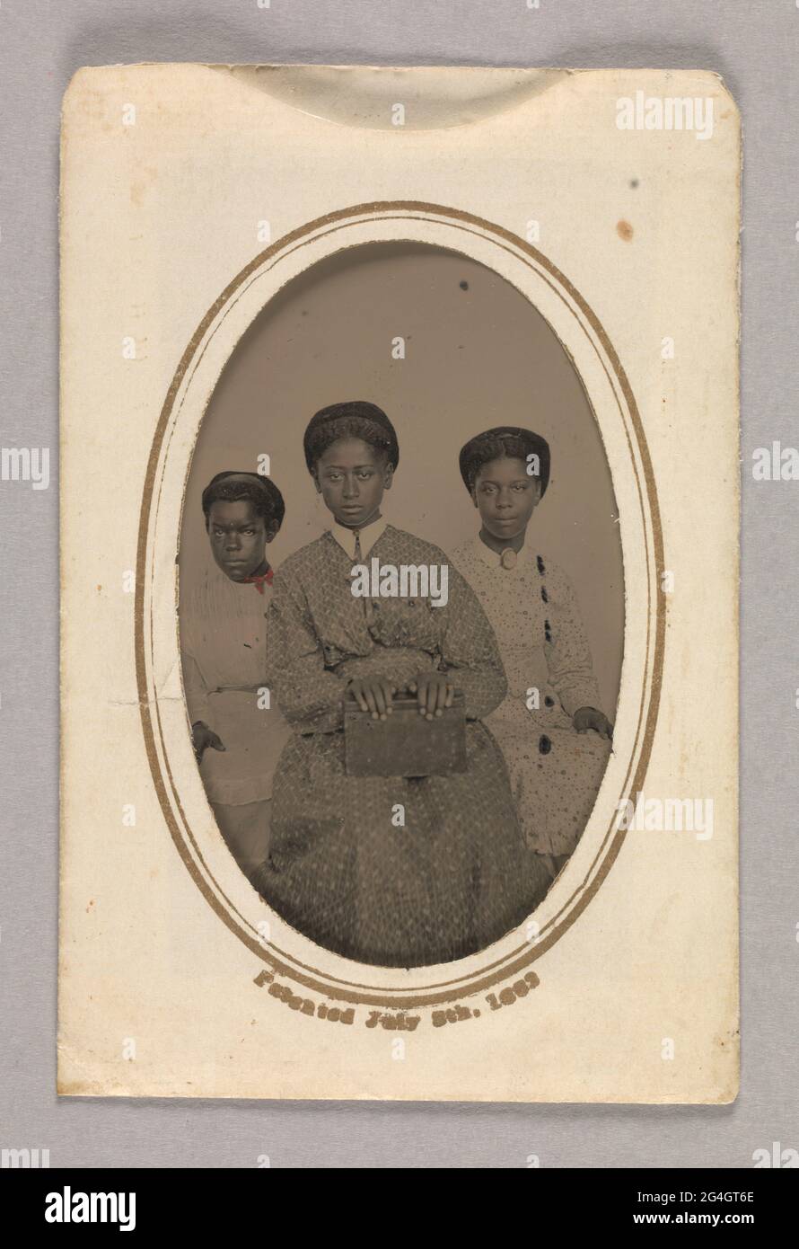 Dieser Tintype (A) zeigt drei (3) nicht identifizierte junge afroamerikanische Frauen, die sitzen und der Kamera gegenüberstehen. Die Frau in der Mitte steht etwas vor den beiden anderen. Sie trägt ein dunkelfarbenes Kleid mit einem helleren Aufdruck und einem spitzen weißen Kragen. Sie hält eine Schachtel oder ein dickes Buch in ihrem Schoß, und vergoldet wird einem ihrer linken Finger und in der Mitte ihres Kragens hinzugefügt. Die Frau auf der rechten Seite trägt ein helles Überkleid mit dunklem Blumendruck und einen hellen Rock mit gerafftem Saum. Vergoldet wird eine ovale Brosche, die sie an ihrem Hals über einem kleinen, runden Kragen trägt. Der Stockfoto