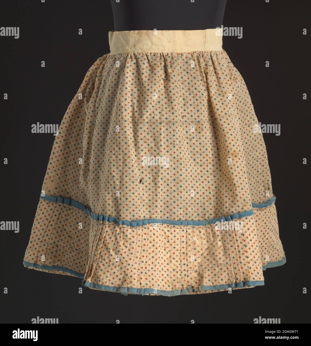 Ein runder Kinderrock im Blumenmuster mit Faltenmuster am Saum. Der Hauptstoff ist ein einfarbiger Naturfaserstoff mit cremefarbenem Boden und einem sich wiederholenden Muster aus kleinen vierblättrigen Blüten in Rot, Lila, Blau und Bräune. Der Rock ist überwiegend handgenäht. Er hat einen Bund aus ungefärbtem Bastfaser-Stoff mit dem Blumendruck, der gerafft und am Bund mit einer Peitsche genäht ist. Der Rock ist auf dem Rücken enger gerafft. Es gibt einen Schlitz in der Mitte hinten, der den Bund vollständig teilt. Es gibt keine angebrachte Verschlussmethode, aber Rostfärbung an der hinteren Öffnung des Stockfoto