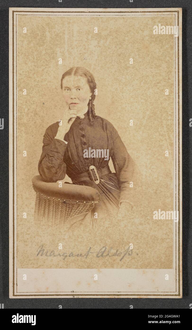 Carte-de-Visite von Margaret Alsop im Halbportrait. Alsop sitzt mit ihrem rechten Ellenbogen auf dem Rücken eines Stuhls und ihrer rechten Hand, die ihr Kinn berührt. Sie schaut direkt auf die Kamera. Alsop trägt ein dunkelfarbenes, hochhalsiges Kleid mit Knöpfen vom Hals bis zur Taille und einem Gürtel mit ovaler Schnalle, die die gefaltete Taille einschnürt. Am Kragen und an den Ärmelabschlüssen ist eine helle Zierleiste vorhanden. Das Foto hat Retusche in Alsops Augen und rosa Handfärbung auf ihren Lippen und Wangen.;Es gibt eine Inschrift in Graphit handgeschrieben am unteren Rand des Drucks, die &q lautet Stockfoto