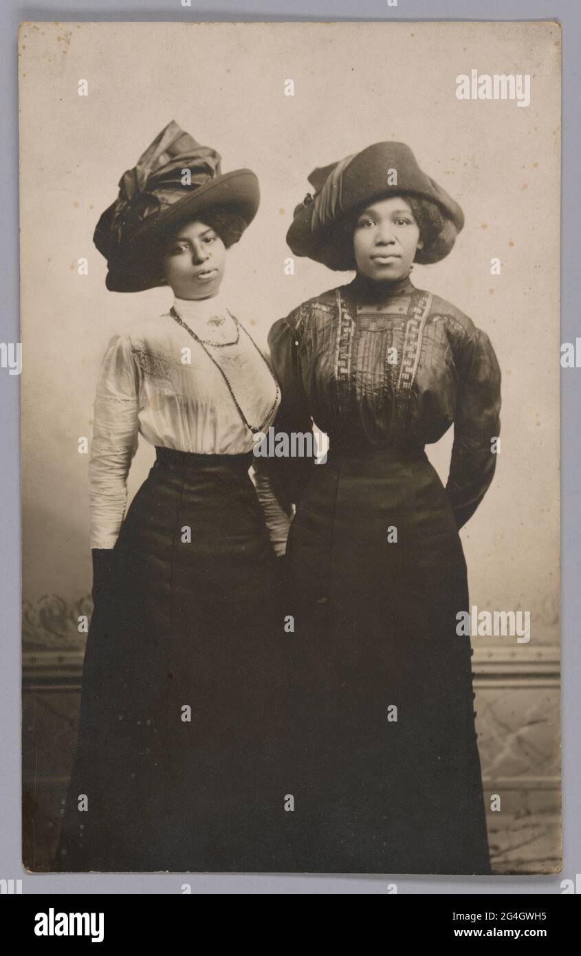 Das lange Porträt zeigt die beiden Frauen, die nebeneinander stehen und große Hüte mit langärmeligen Blusen und langen Röcken tragen. Sie schauen mit den Händen hinter dem Rücken auf die Kamera. Stockfoto
