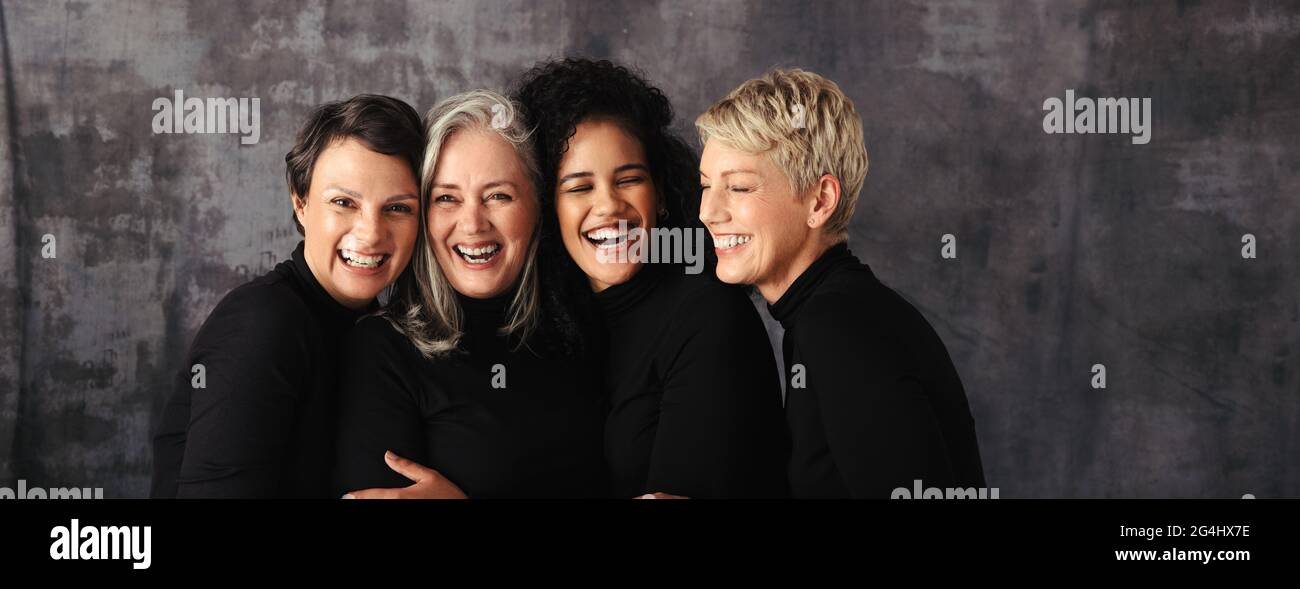 Lächelnde Frauen unterschiedlichen Alters stehen im Studio zusammen. Eine Gruppe von vier selbstbewussten Frauen, die ihre natürlichen und alternden Körper gegen ein Studio umarmen Stockfoto