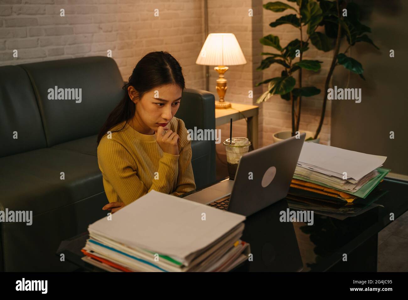 Müde junge asiatische Geschäftsfrau, die mit Händen am Kinn denkt, während sie in der späten Nacht zu Hause auf den Laptop-Bildschirm starrt, auf dem sich Arbeitsdateien im Büro auf dem Tisch befinden Stockfoto