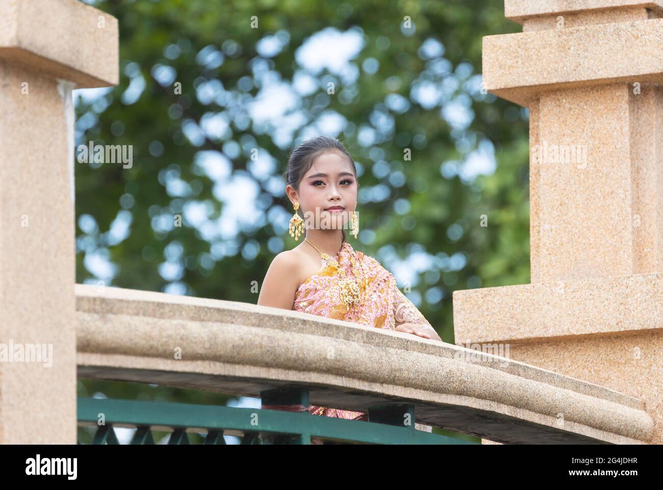 Ein asiatisches Schulmädchen in traditioneller thailändischer Kleidung steht am Geländer einer Brücke in einem Park. Stockfoto