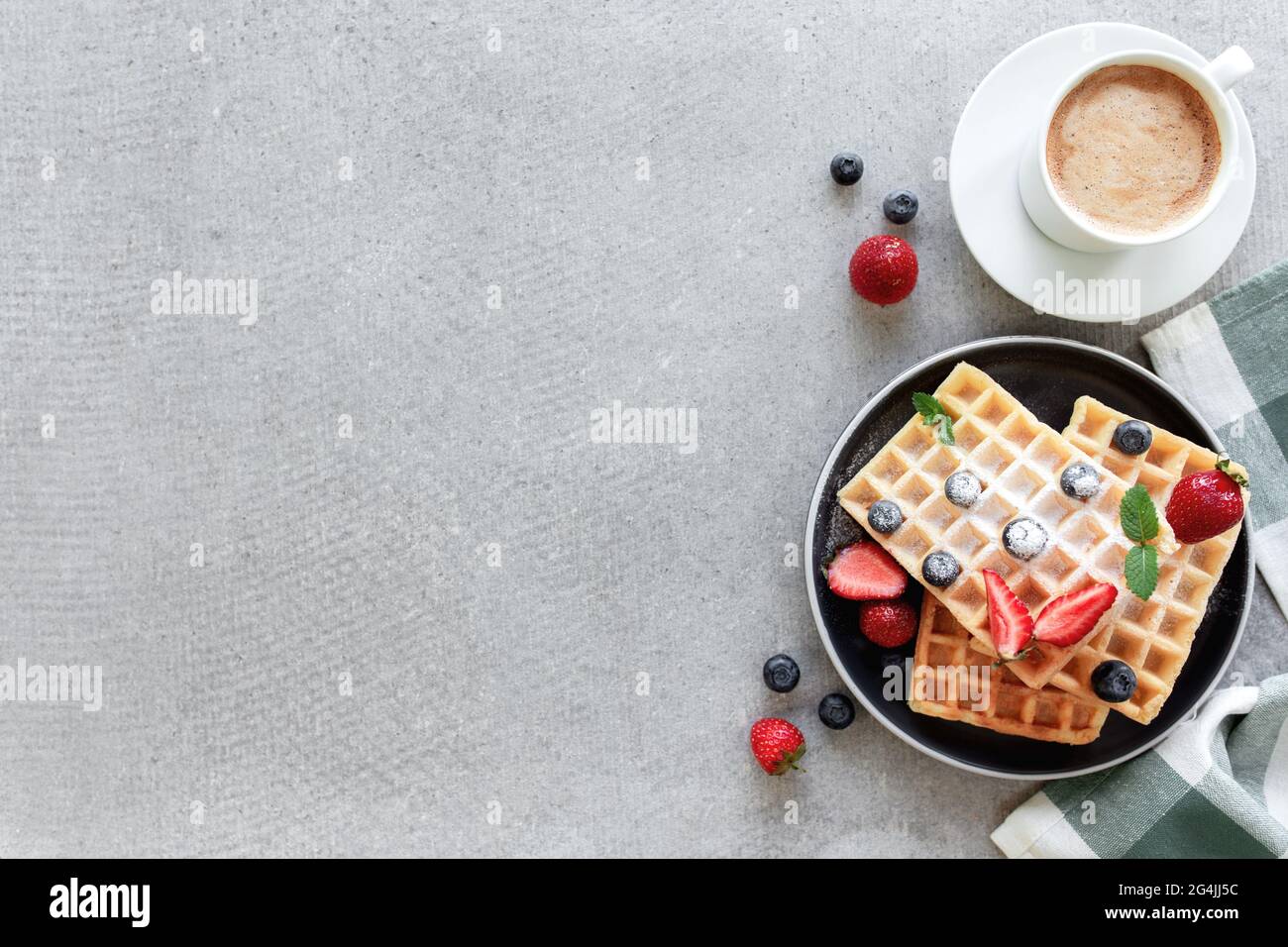 Stapel von zuckerpulverförmigen Waffeln mit Honig auf einem Teller auf dem Handtuch und grauen Beton- oder Schiefertisch mit Heidelbeere, gehackten Erdbeeren und Minzblättern Stockfoto