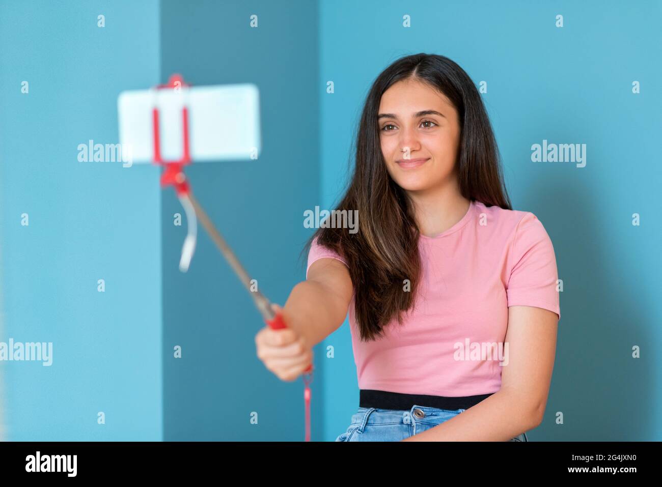 Hübsches Teenager-Mädchen lächelt, während sie Selfie-Bilder mit ihrem Handy für soziale Medien und Online-Kommunikation gegen blaue Wand für Kopie sp Stockfoto