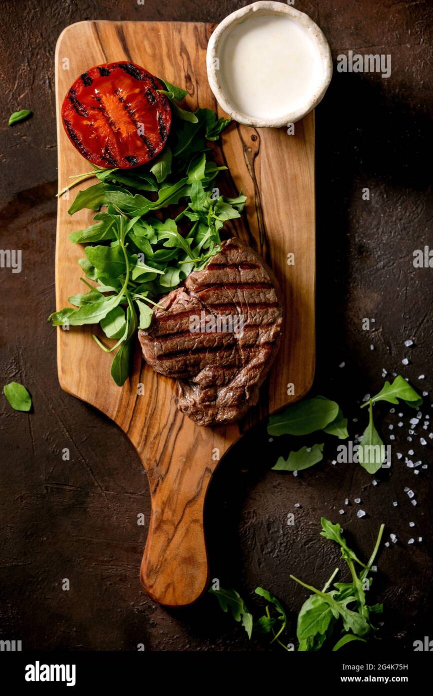 Gegrilltes Rindersteak serviert mit cremiger Sauce, gegrillter Gemüsestomate und Rucola-Salat auf Holzschneidebrett auf dunkelbraunem Hintergrund. T Stockfoto
