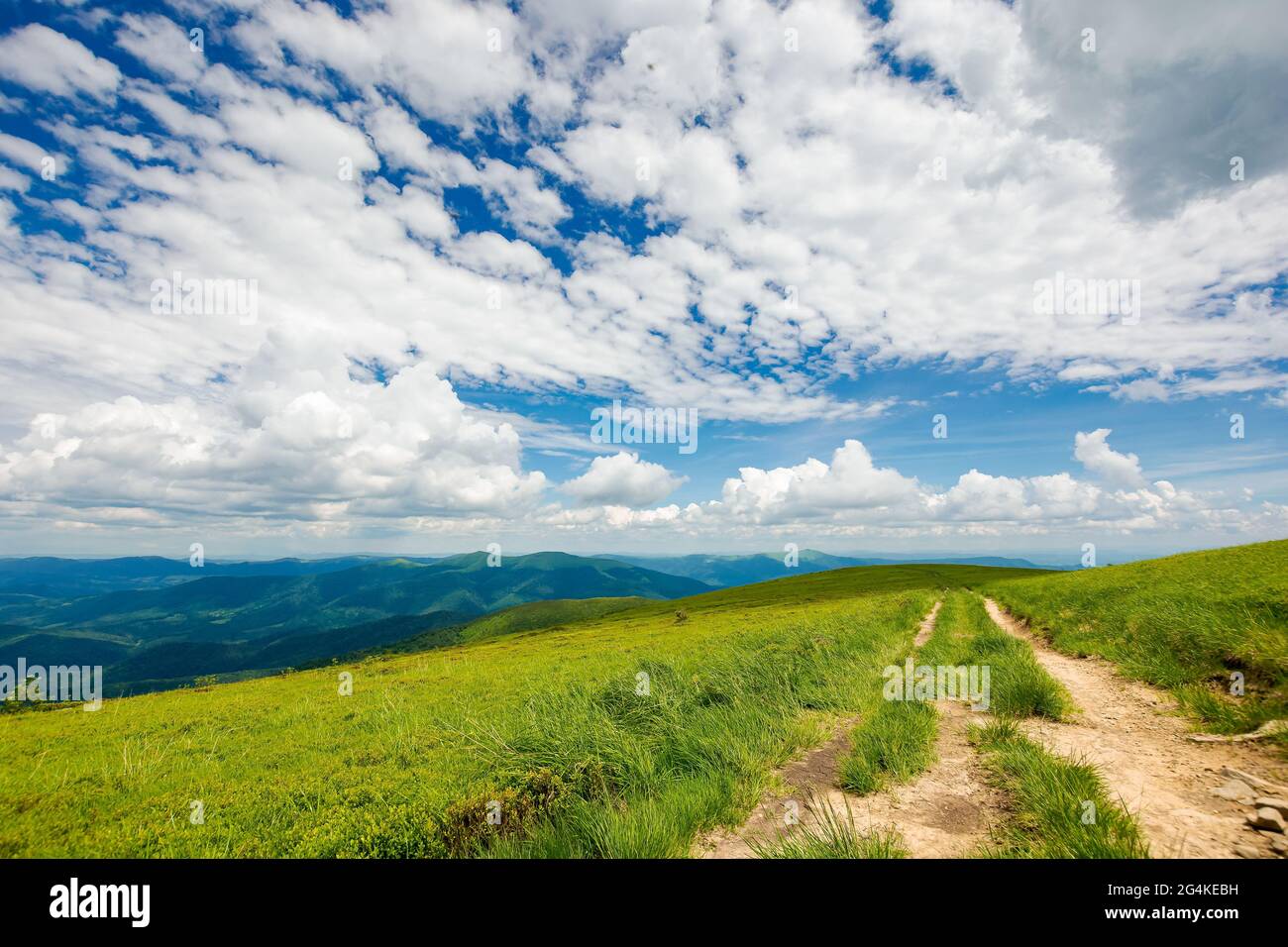 Landstraße durch grasbewachsene Hügel. Bergrücken in der Ferne unter einer wunderschönen Wolkenlandschaft am blauen Himmel. Reise-Backcountry-Konzept Stockfoto