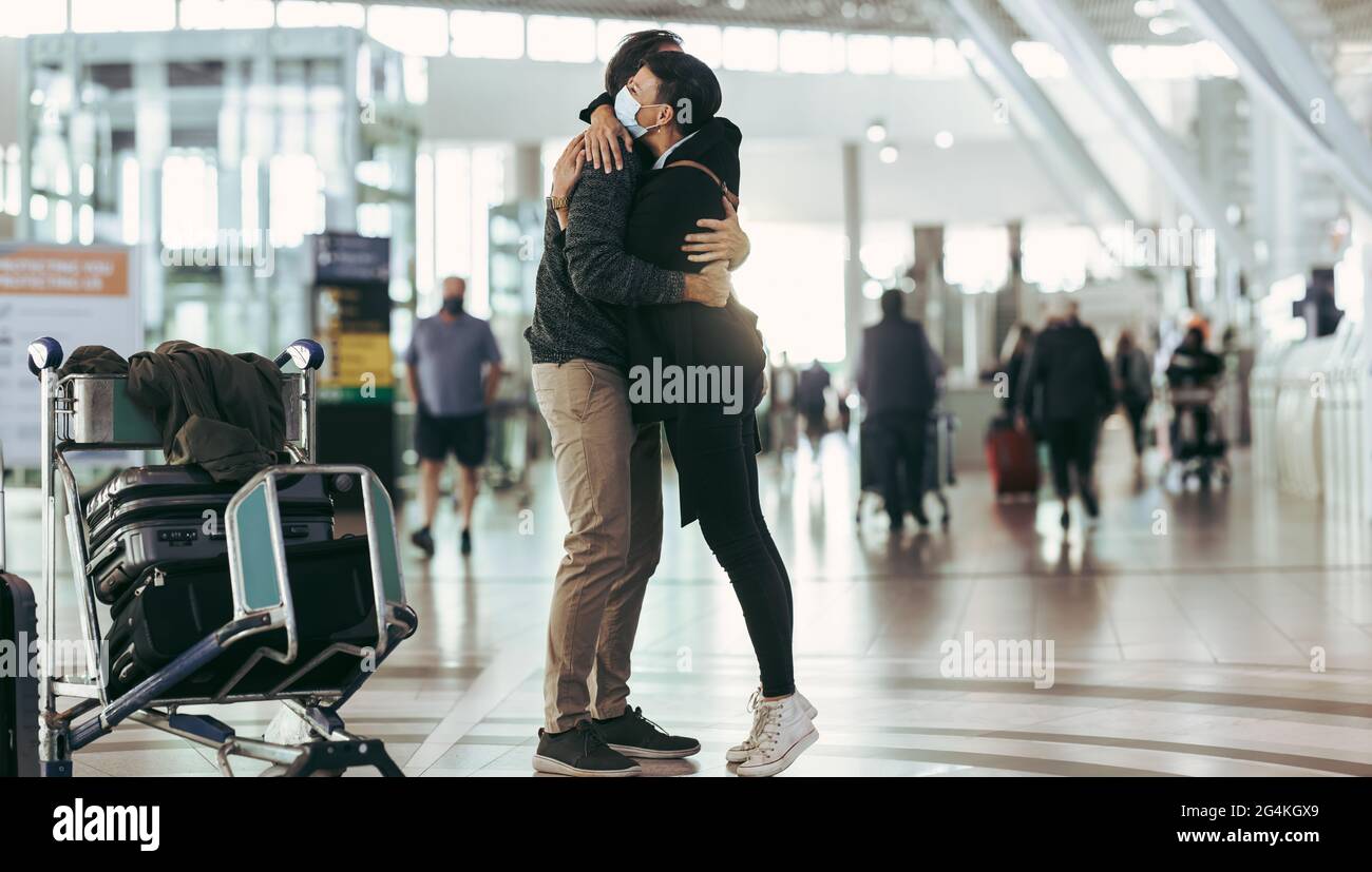 Frau in Gesichtsmaske, die einen Mann bei der Ankunft am Flughafen empfängt. Das Paar trifft sich am Ankunftseingang des Flughafens und umarmt sich. Stockfoto