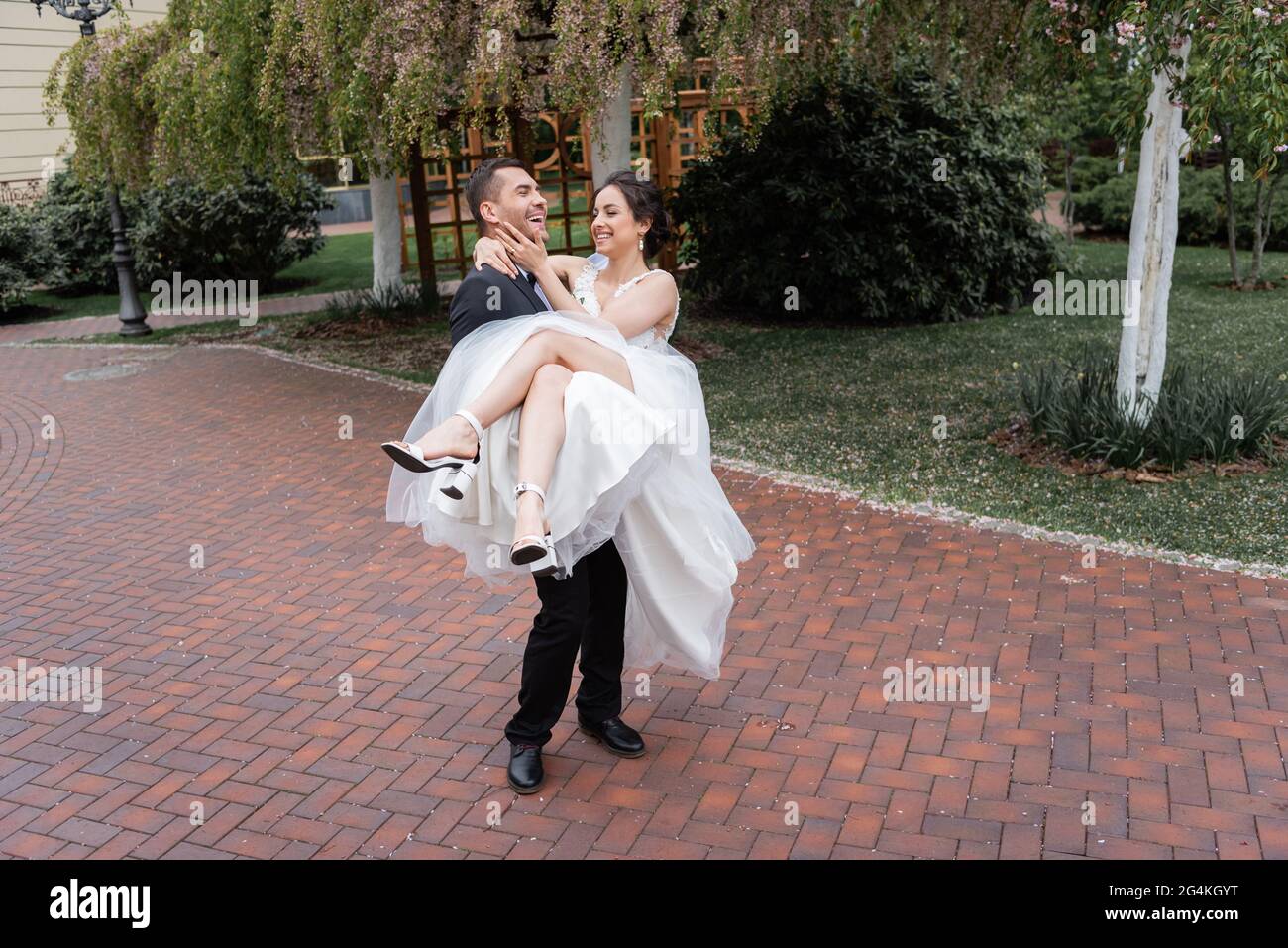 Fröhliche Braut im Schleier und Hochzeitskleid berühren Gesicht des Bräutigams im Park Stockfoto
