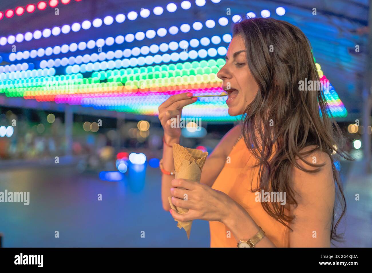 Nahaufnahme einer glücklichen, unbeschwerten jungen Frau, die mit geschlossenen Augen einen Bissen Eis vor verschwommenen, farbenfrohen Karnevalslichtern nimmt Stockfoto