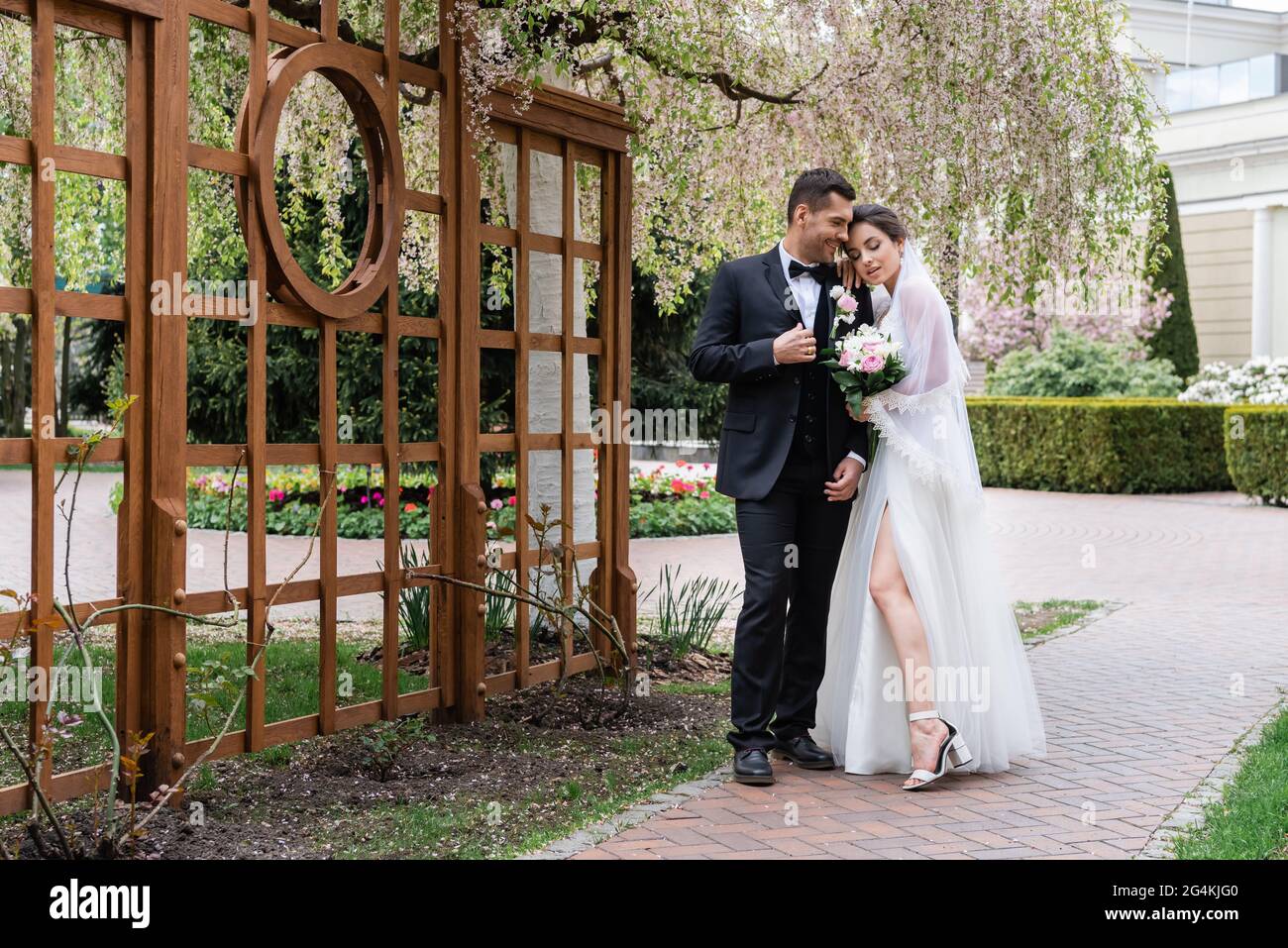 Fröhlicher Bräutigam, der in der Nähe der Braut im Hochzeitskleid und im blühenden Baum im Park steht Stockfoto