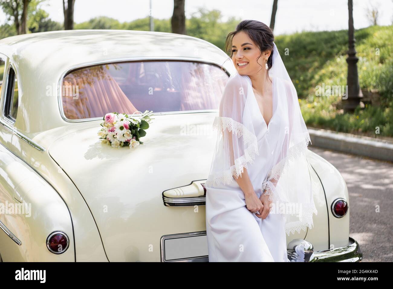 Glückliche Braut im Hochzeitskleid steht in der Nähe Blumenstrauß auf Retro-Auto Stockfoto