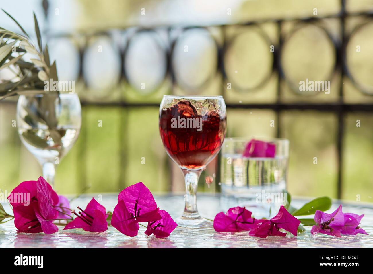 Elegantes helles Glas Rotwein, Wein spritzt ins Glas. Sommercocktails im Hintergrund. Dekorative rosa Blüten und ein Zweig des Olivenbaums Stockfoto