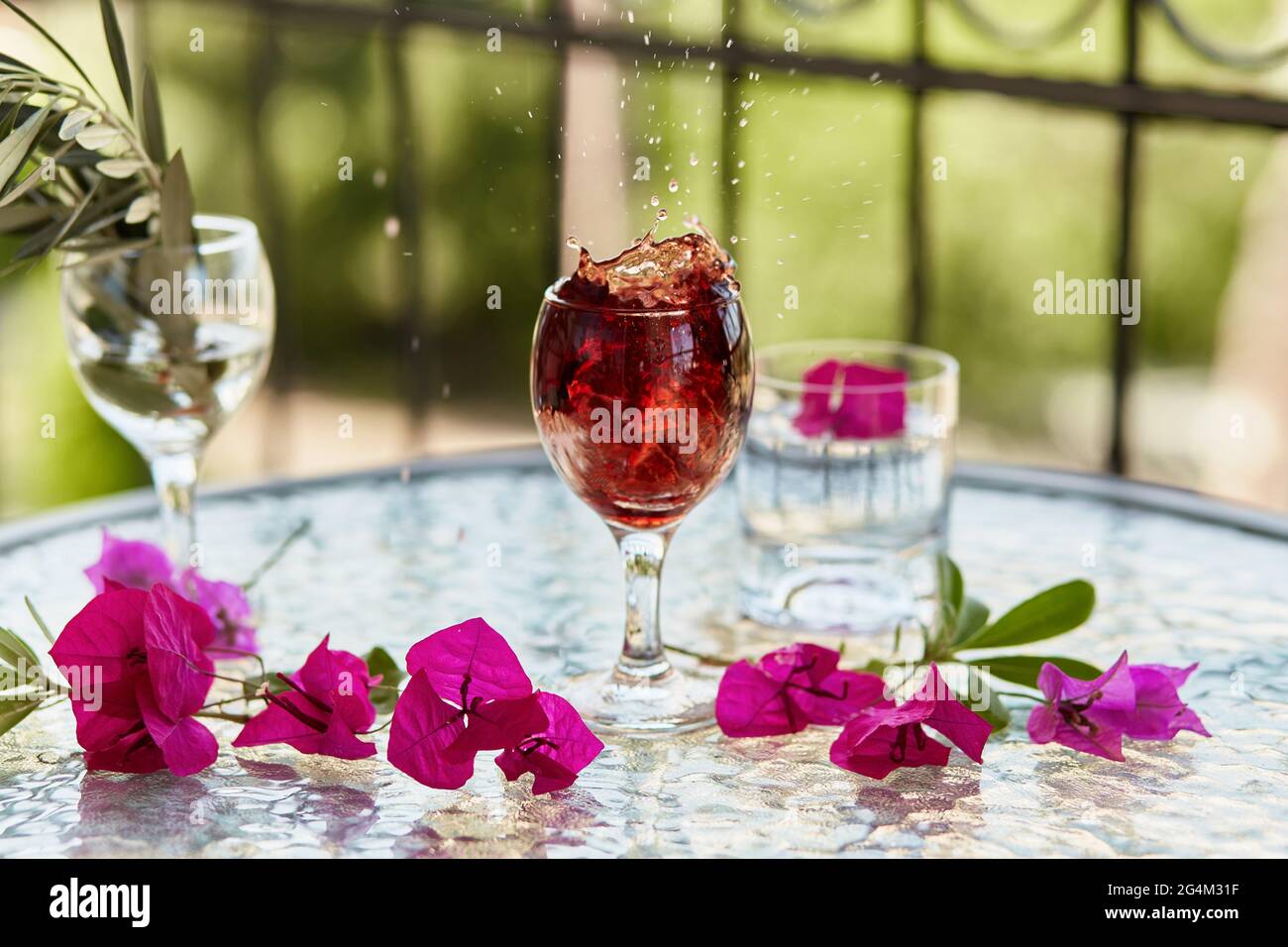 Elegante und farbenfrohe Explosion von Rotwein im Glas. Im Sommer helle Cocktails auf dem Hintergrund. Dekorative rosa Blumen und der Zweig des olivgrünen t Stockfoto