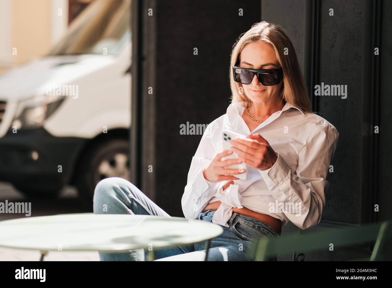 Elegante kaukasische Frau, die an einem Cafe-Tisch sitzt und auf dem Smartphone textet, selbstbewusste Unternehmerin, die eine Sonnenbrille und ein weißes Hemd trägt Stockfoto