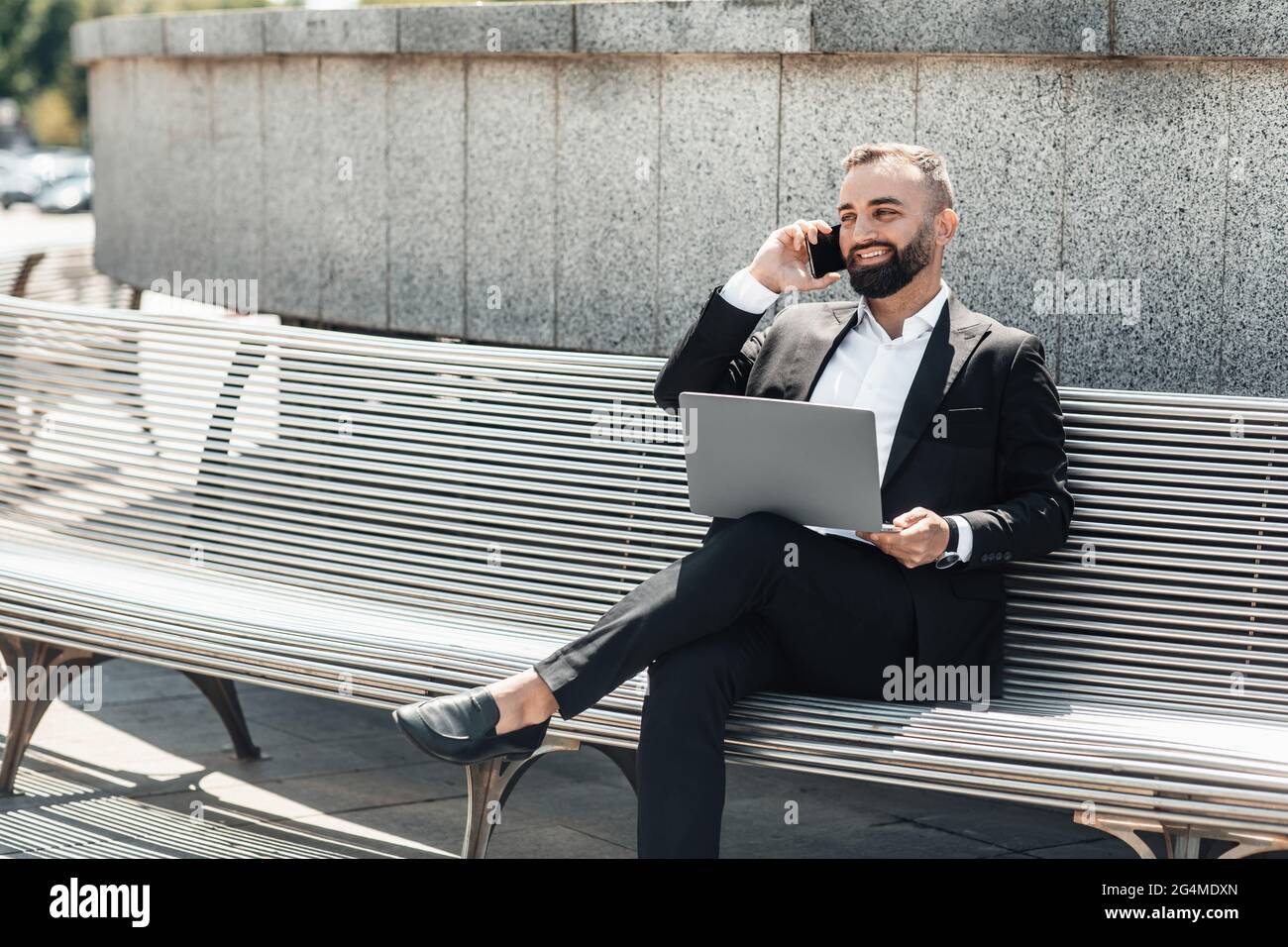 Reifer bärtiger Geschäftsmann mit modernem Laptop, der auf einer Bank sitzt und auf dem Smartphone redet, leerer Raum Stockfoto