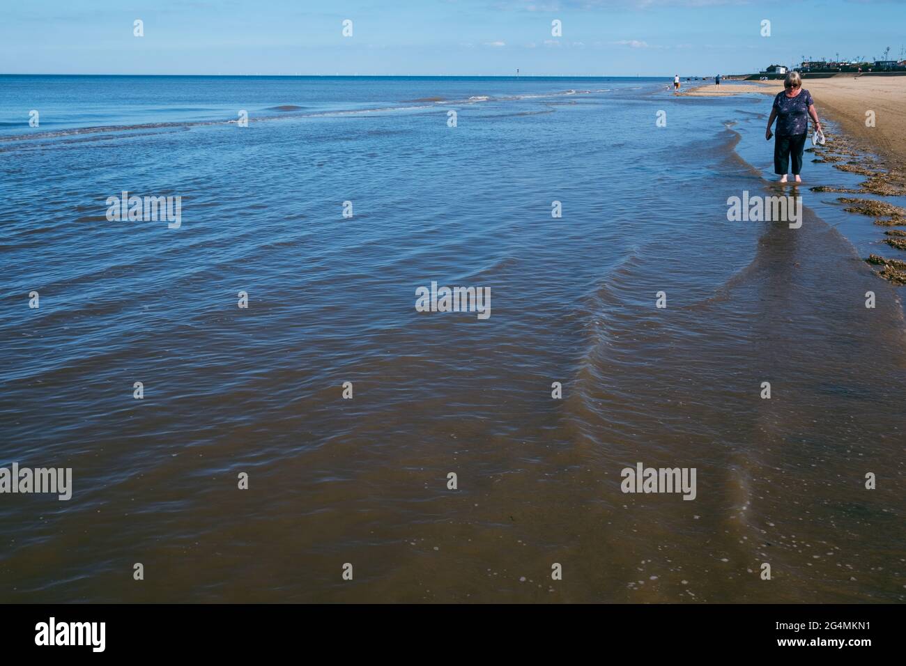 Eine blonde Frau mittleren Alters schlenderte barfuß im Meer entlang der Küste des North Beach von Mablethorpe in Lincolnshire Stockfoto