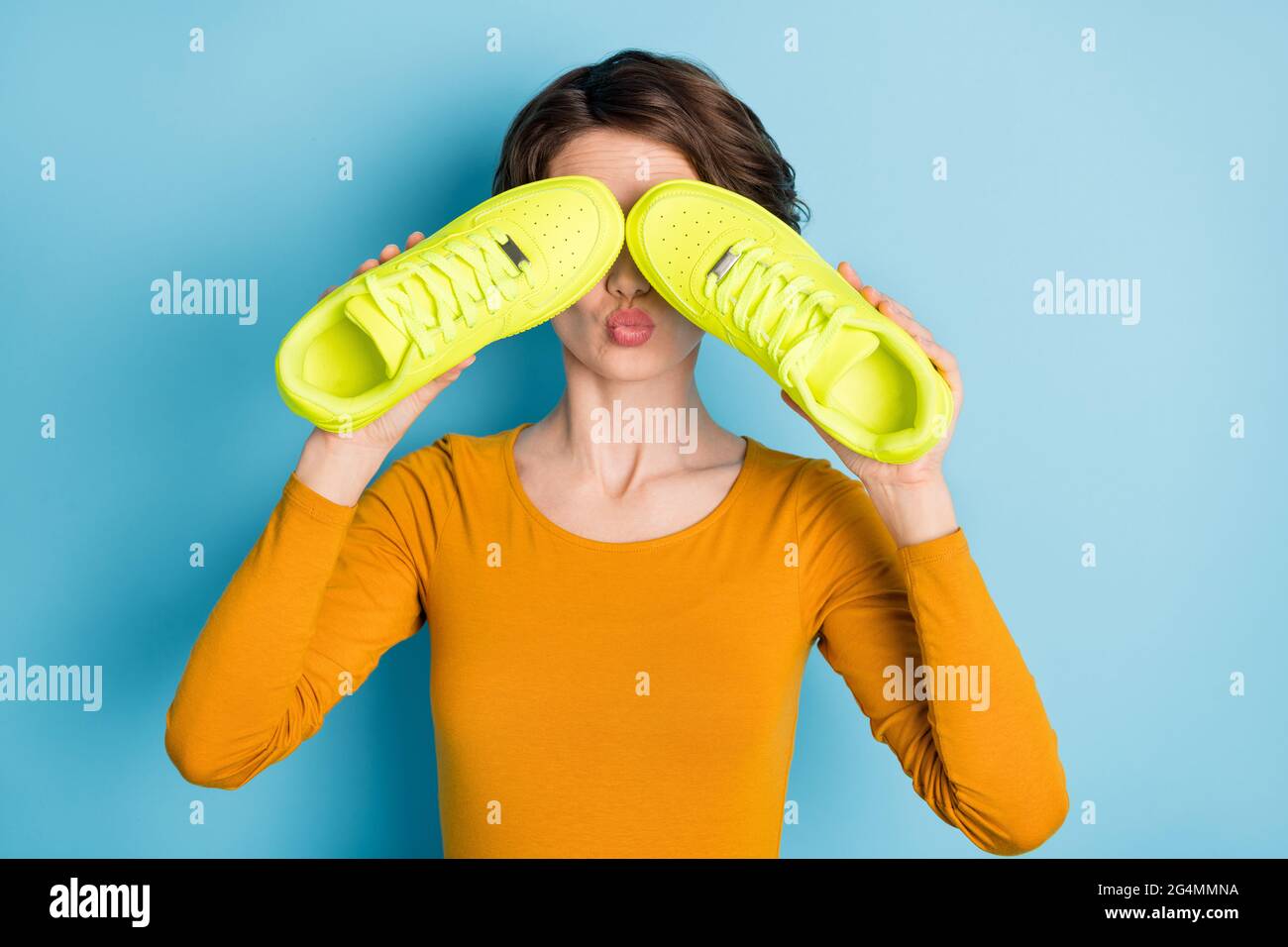 Foto von schönen flirty Brünette Dame Cover Gesicht von Turnschuhen tragen gelbes Hemd isoliert in lebendigen blauen Hintergrund Stockfoto