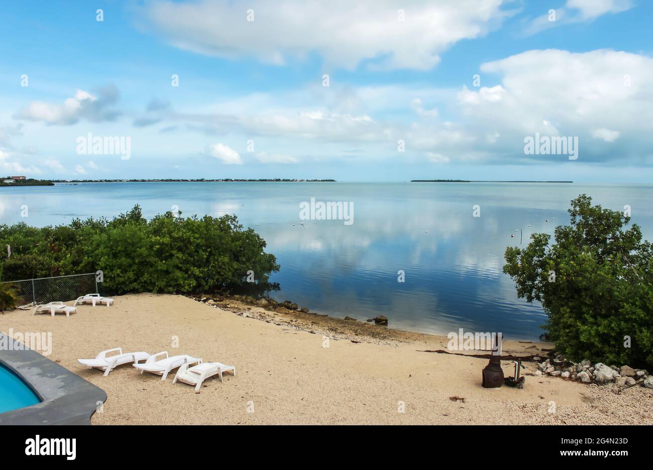 Blick auf den Strand mit Liegestühlen und Chiminea am Rand des stillen Ozeans mit Spiegelung des Himmels und Swimmingpool am Rand Stockfoto