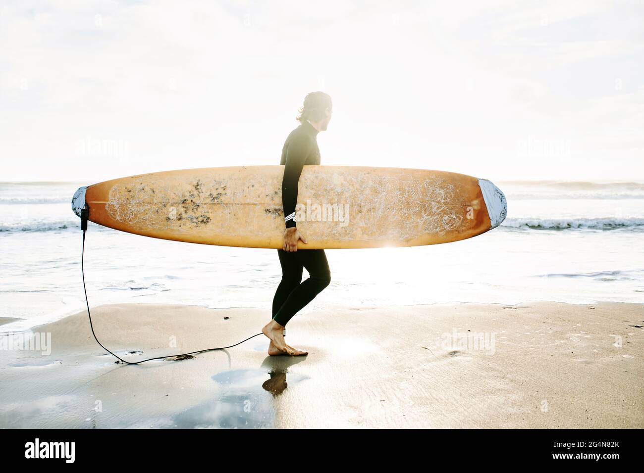 Seitenansicht des in Neoprenanzug gekleideten Surfers, der am Morgen mit dem Surfbrett am Strand spazierenging, während der Sonnenaufgang im Hintergrund zu beobachten war Stockfoto