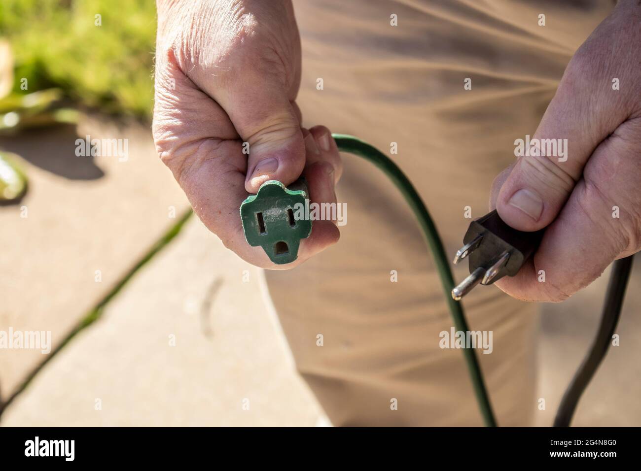Nahaufnahme der Hände eines Mannes, die sich fertig machen, um das amerikanische Netzkabel mit drei Zapfen in das abgenutzte grüne Verlängerungskabel einzustecken Stockfoto