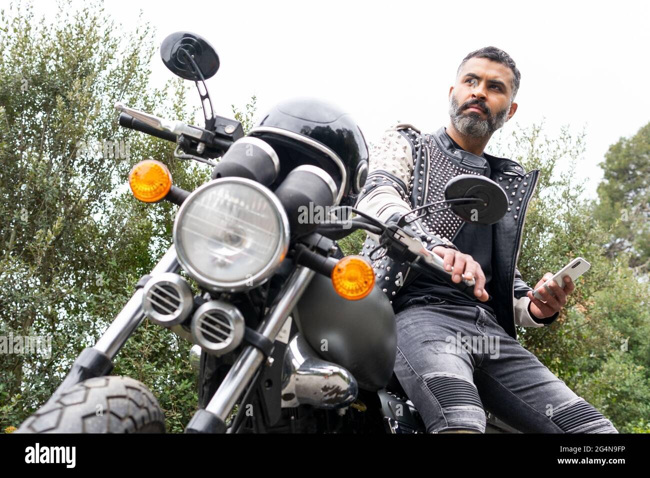 Niedriger Winkel der konzentrierten Erwachsenen brutalen ethnischen männlichen Biker mit Bart in Lederjacke mit Nieten sitzen auf modernen Motorrad und wegschauen, während m Stockfoto