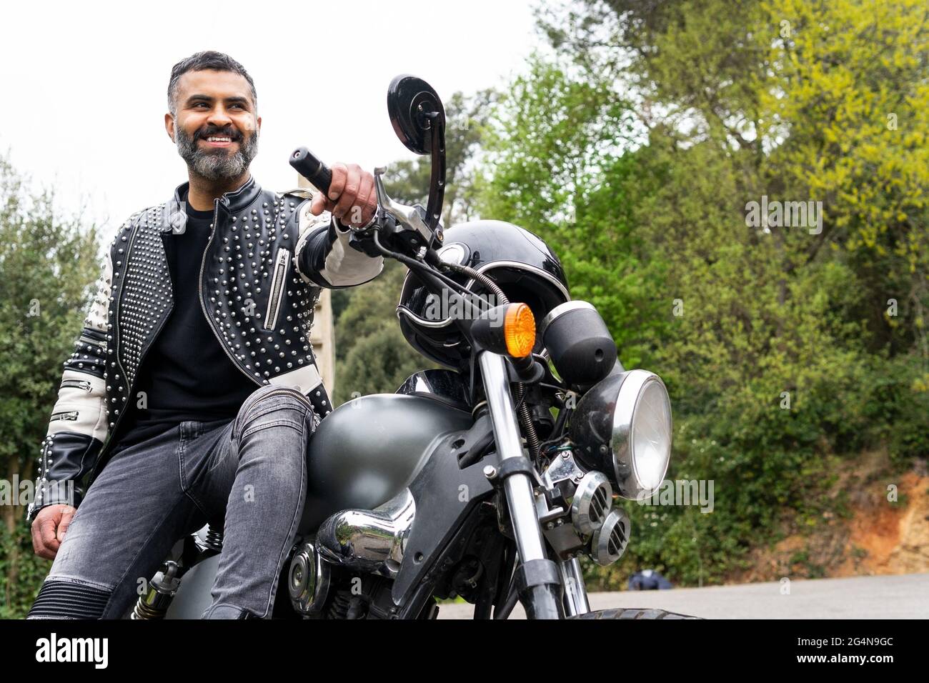Von unten bärtiger, ethnischer Biker in schwarzer Lederjacke, der auf einem modernen Motorrad auf einer Asphaltstraße inmitten üppig grüner Bäume in den Bergen sitzt Stockfoto