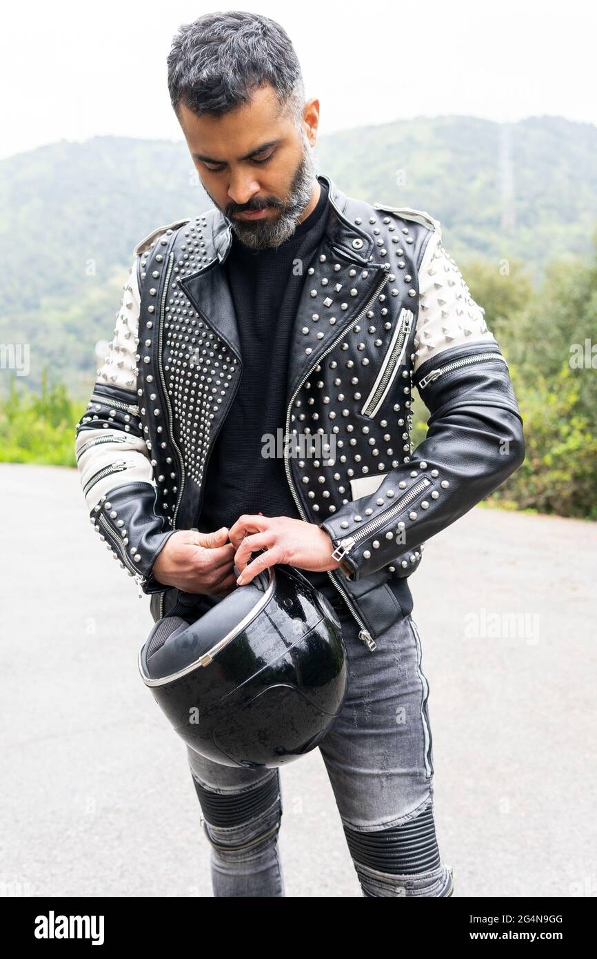 Serious brutalen ethnischen männlichen Biker in stilvoller Lederjacke auf Asphalt Straße mit Helm in der Hand in der Nähe von üppigen grünen Bäumen wächst in bergigen V Stockfoto
