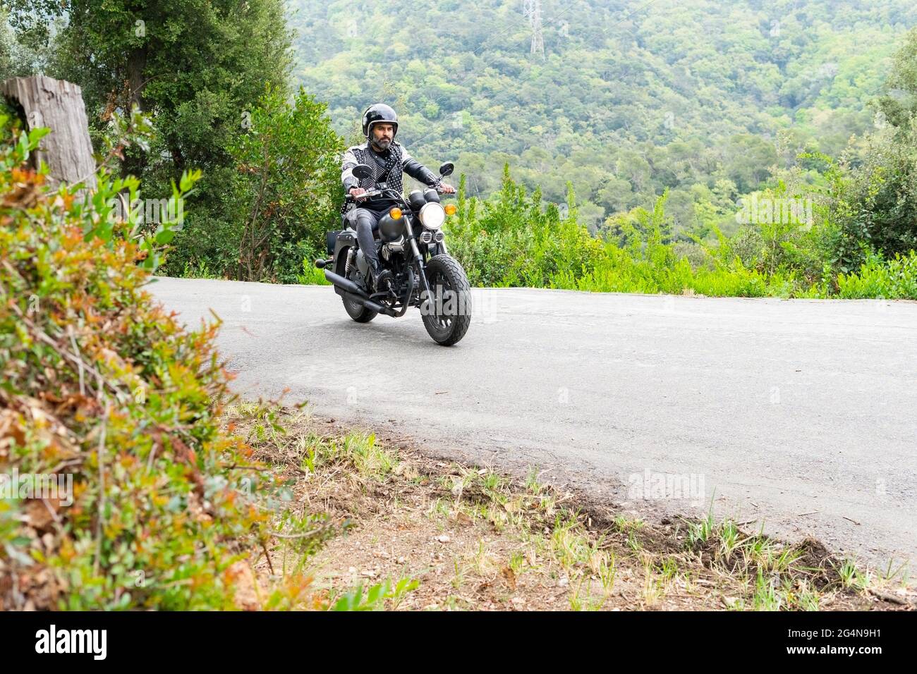 Voller Körper fokussierter bärtiger ethnischer Biker in schwarzer Lederjacke und Helm, der auf einem modernen Motorrad auf einer Asphaltstraße inmitten üppig grüner Bäume unterwegs ist, wächst Stockfoto