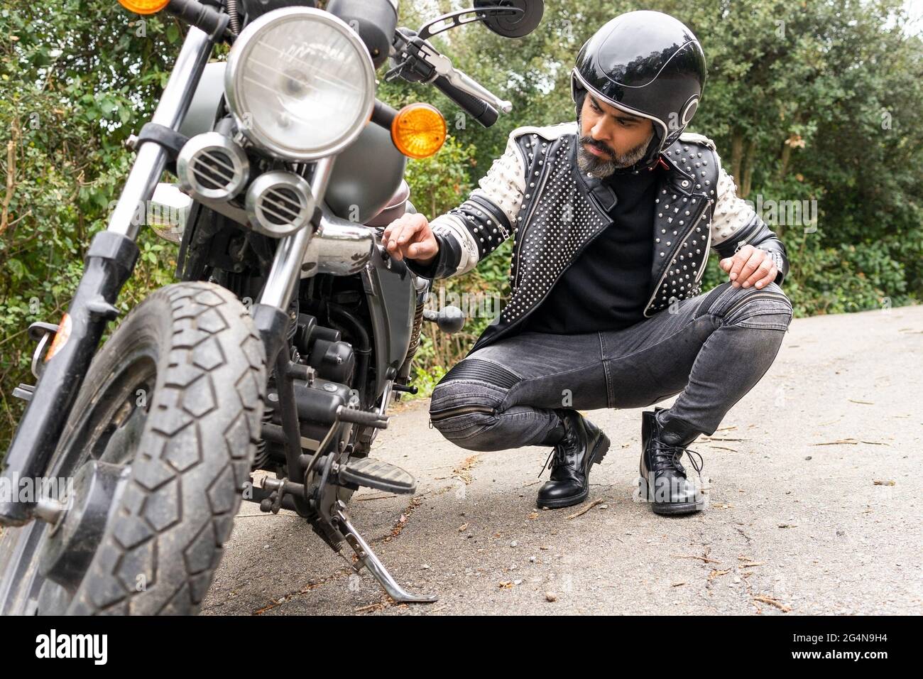 Ganzer Körper von ernsthaften erwachsenen ethnischen männlichen Biker in stilvollem Outfit und Helm sitzen auf Haunches und Überprüfung gebrochenen Motorrad Details auf Straße in der Nähe von Lush Stockfoto
