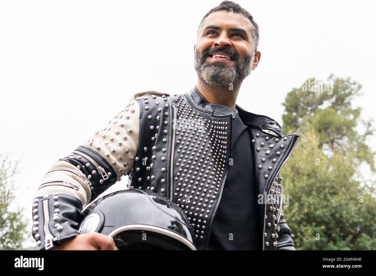 Von unten brutal ethnischen männlichen Biker in stilvoller Lederjacke auf Asphalt Straße mit Helm in der Hand in der Nähe von üppigen grünen Bäumen wachsen in Mountainou Stockfoto