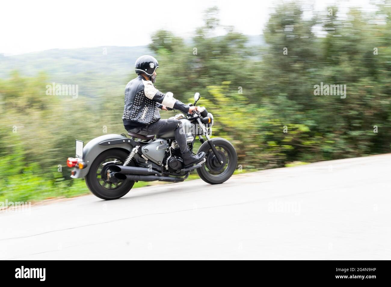 Ganzer Körper von bärtigen ethnischen männlichen Biker in schwarzer Lederjacke und Helm Reiten moderne Motorrad auf Asphaltstraße inmitten üppig grünen Bäumen in m wachsen Stockfoto