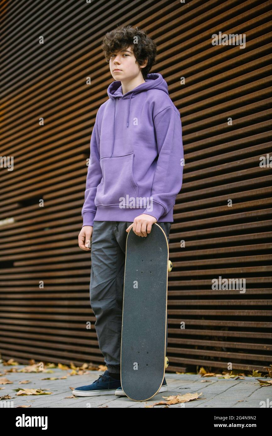 Ganzer Körper eines ernsthaften jungen Mannes in legerer Kleidung mit  Skateboard, das die Kamera anschaut, während es in der Nähe einer Wand auf  der Straße steht Stockfotografie - Alamy