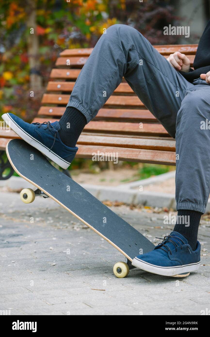 Nicht erkennbarer Mann in Sneakers balanciert auf Skateboard, während er auf einer Holzbank im Park mit Büschen während des Trainings in der Stadt sitzt Stockfoto